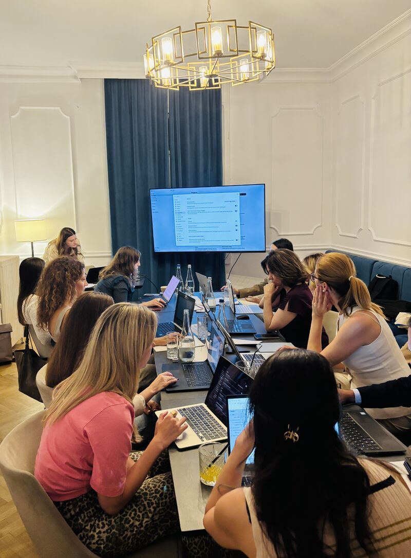 AI Circle members with laptops open around a long table at A46, Claude on the TV screen